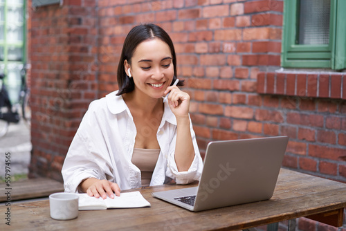 Young smiling asian woman with wireless earphones, sitting in coworking office space with laptop, doing homework, making notes photo