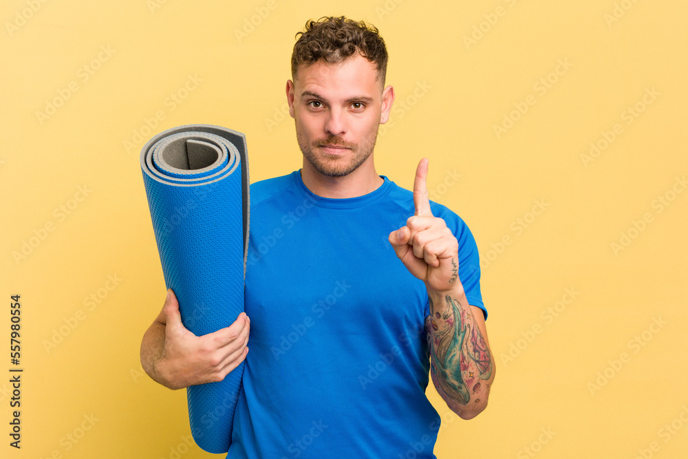 Φωτογραφία Young caucasian sport man holding a mat isolated on yellow background showing number one with finger
