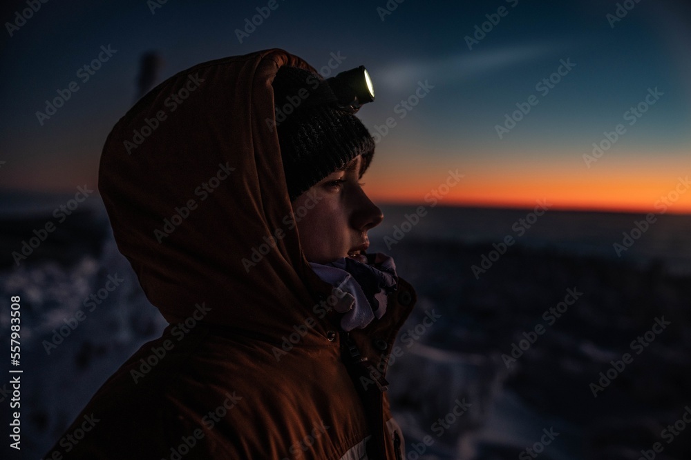 Teenager boy in a warm winter jacket with a headlamp on a mountain winter trail. Minutes before sunset, Babia Gora, Beskids, Poland - obrazy, fototapety, plakaty 