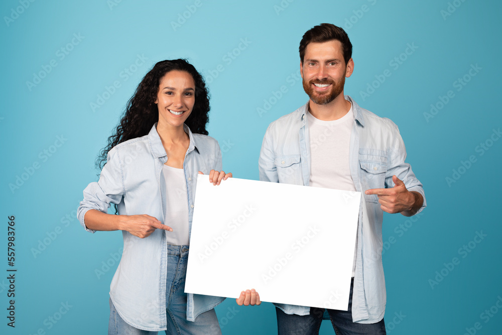 Happy young middle eastern wife and european bearded husband show finger on banner with blank space Happy young middle eastern wife and european bearded husband show finger on banner with blank space