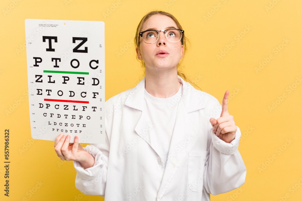 Young caucasian oculist woman holding a eye chart paper isolated on yellow background pointing upside with opened mouth. Young caucasian oculist woman holding a eye chart paper isolated on yellow background pointing upside with opened mouth.