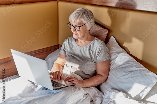 Senior smiling woman with short gray hair, using laptop in bed.