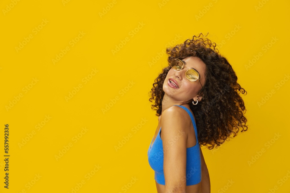 Woman with curly afro hair in a blue T-shirt on a yellow background dancing flying hair with sunglasses yellow, hand signs, look into the camera, smile with teeth and happiness, copy space