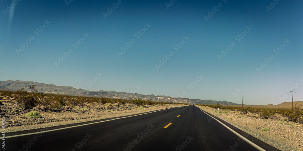 Straight road in small desert plants and sand with mountains in america at sunny day