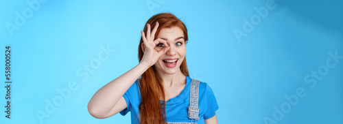 Cheerful upbeat redhead european female feel lucky inspired, look through circle okay on eye, smiling broadly amused, look wondered admire cool awesome sales, stand blue background