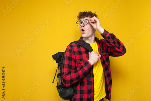 curly guy student in glasses with a backpack stands on a yellow background and looks back at the copyspace in surprise photo