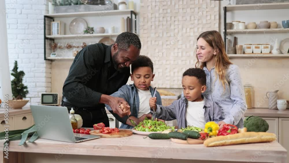 Young happy mom, dad and little sons adding olive oil to the salad while cooking in kitchen together. Happy multiracial parents and small boys kids have fun preparing healthy food at home. Young happy mom, dad and little sons adding olive oil to the salad while cooking in kitchen together. Happy multiracial parents and small boys kids have fun preparing healthy food at home.