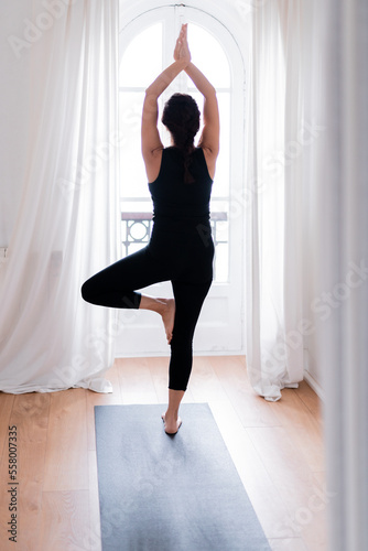 Vertical photo of woman in uniform sportswear top, leggings. Doing yoga asana exercise, meditating at home near window, on a mat. Healthy sport