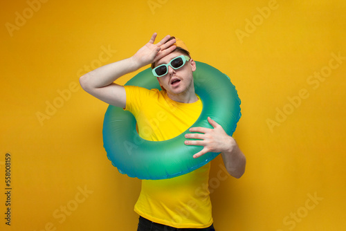 young guy in summer clothes and glasses on vacation with an inflatable ring wipes his forehead with his hand
