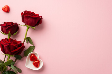 Valentine's Day concept. Top view photo of bouquet of red roses and heart shaped saucer with chocolate candies on isolated pastel pink background with copyspace