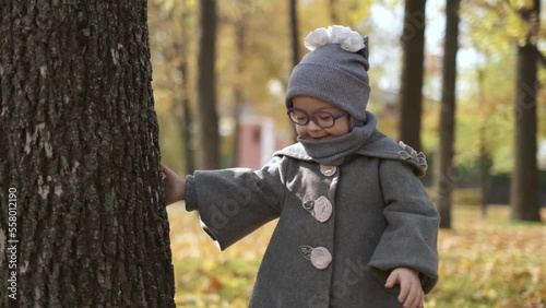 Cute little girl with health disorder wearing warm coat and hat and stylish eyeglasses peeking out from behind a tree, playing peek-a-boo, having fun enjoying warm weather in bright autumn park