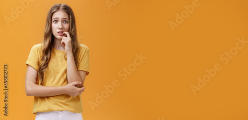 Girl worried mom react on new tattoo. Portrait of anxious and nervous silly insecure young female with wavy natural hairstyle biting finger panicking looking concerned and troubled over orange wall