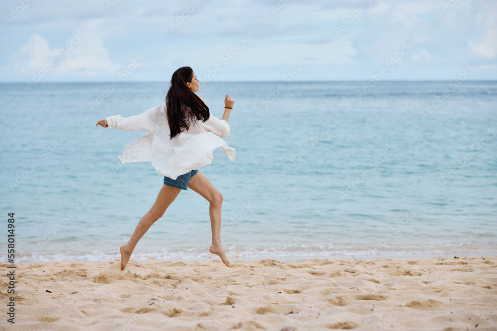 Sports woman runs along the beach in summer clothes on the sand in a yellow tank top and denim shorts white shirt flying hair ocean view, beach vacation and travel Sports woman runs along the beach in summer clothes on the sand in a yellow tank top and denim shorts white shirt flying hair ocean view, beach vacation and travel