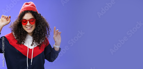 Girl feeling awesome having fun and enoying cool party, dancing with raised hands looking down smiling broadly wearing sunglasses and stylish beanie posing against blue background photo