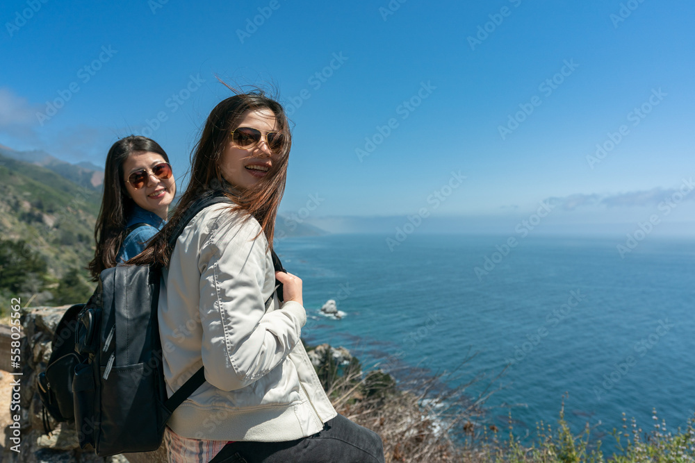 two happy asian Korean female backpacker smiling at camera with blowing hair in winds at a scenic spot overlooking blue sea in big sur usa
