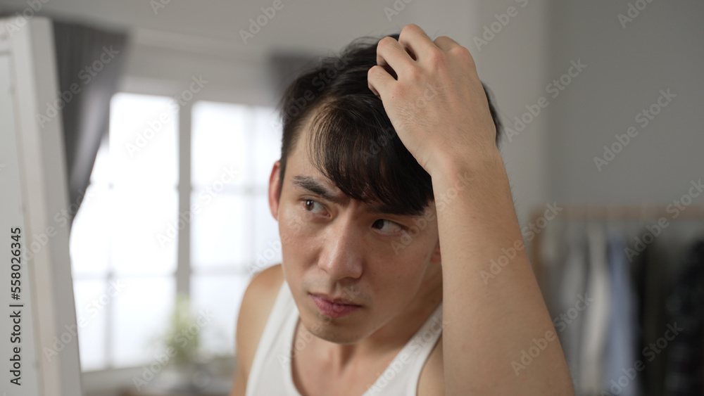 unhappy taiwanese man checking hairstyle in the mirror is shocked and worried about hair loss problem while preparing for a date in the bedroom at home. unhappy taiwanese man checking hairstyle in the mirror is shocked and worried about hair loss problem while preparing for a date in the bedroom at home.