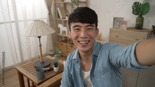 mobile cam view of cheerful asian guy looking and pointing at the camera is talking embarrassedly with hand waving while having a video chat in home living room photo