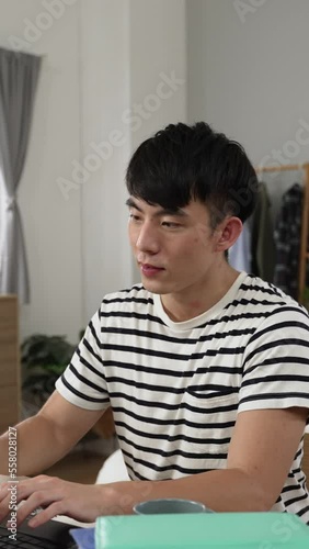 Vertical Screen: Asian Japanese male student learning from home is cheering with hand gesture and taking break with stretch while feeling happy about finishing school project on laptop