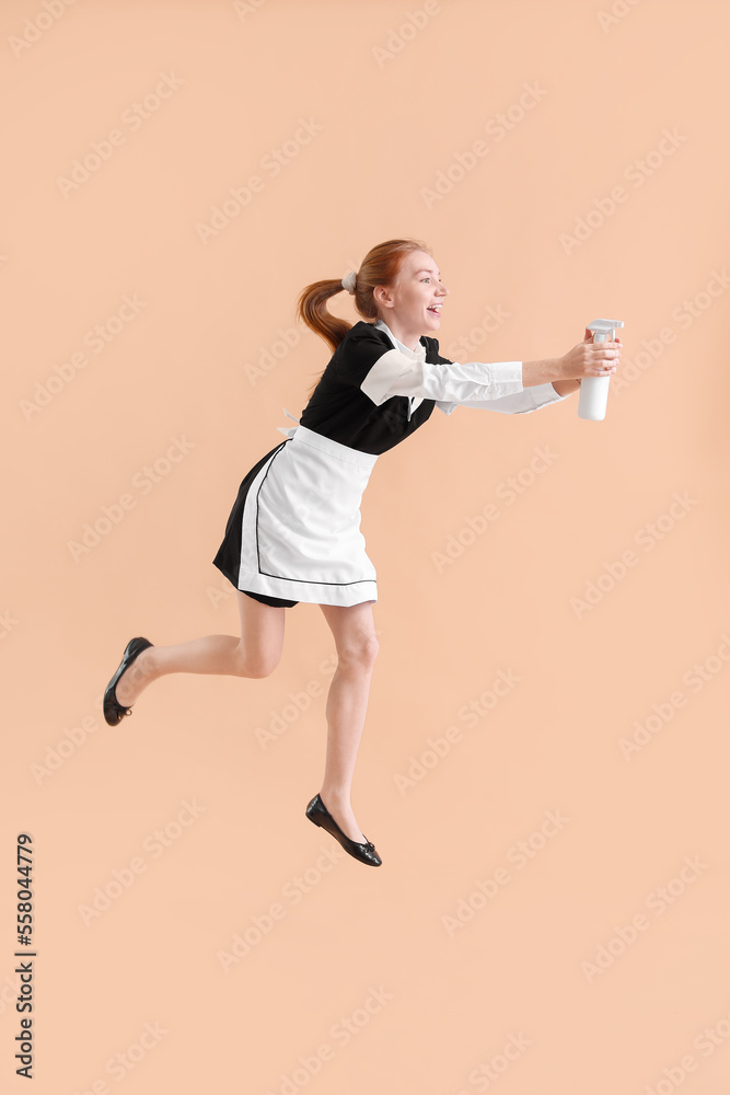 Young female chambermaid with bottle of detergent jumping on beige background