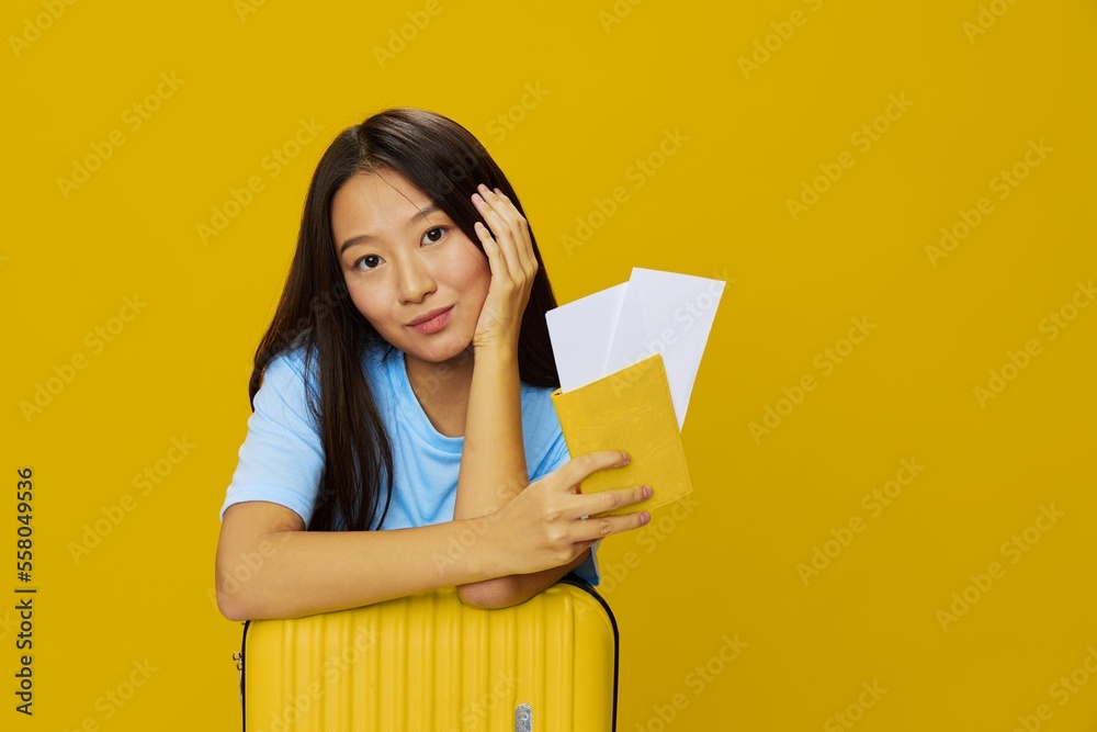 Asian woman traveling with yellow suitcase and tickets with passport in hand, tourist traveling by plane and train with luggage on yellow background in blue T-shirt and jeans Asian woman traveling with yellow suitcase and tickets with passport in hand, tourist traveling by plane and train with luggage on yellow background in blue T-shirt and jeans