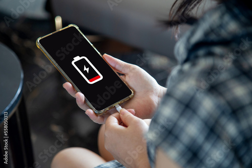 Close up a woman holding a smartphone connected to a charger, displaying a low battery symbol on the screen.