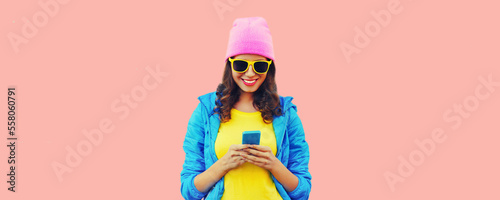 Portrait of stylish happy smiling young woman with smartphone wearing hat, blue jacket on pink background