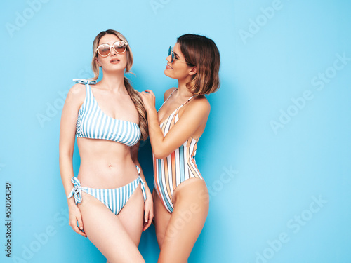 Two beautiful sexy smiling hipster women in summer swimwear bathing suits. Trendy models having fun in studio. Hot female isolated on blue. Cheerful and happy. In sunglasses. Perfect body