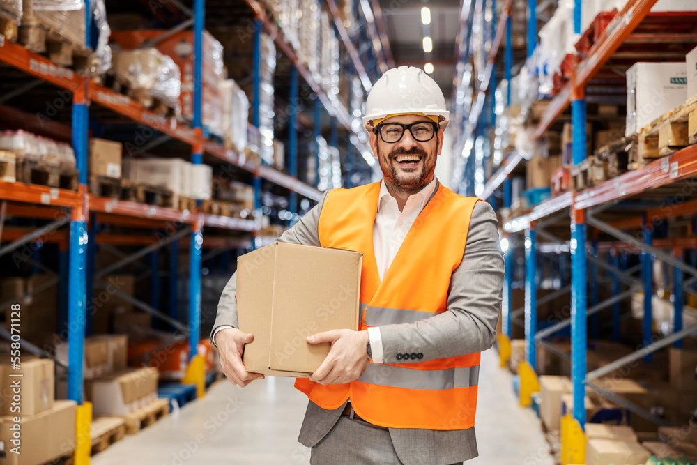 A warehouse manager in suit is holding box with goods and smiling at the camera. A warehouse manager in suit is holding box with goods and smiling at the camera.