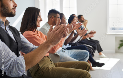 Group of satisfied men and women sitting in row applaud at business conference or team meeting. Side view of multiracial young people sitting in row on chairs in bright hall. Selective focus.