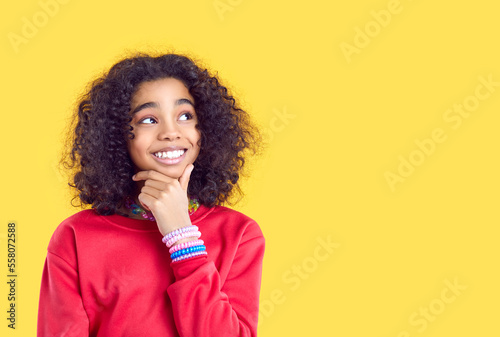 Pensive smiling African American girl isolated on yellow studio background look at copy space aside. Happy biracial teen child consider good sale deal or promotion offer. Advertising concept. photo