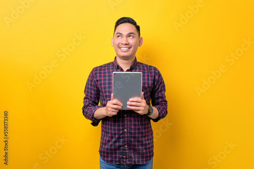Smiling young Asian man holding digital tablet and looking aside at copy space isolated over yellow background