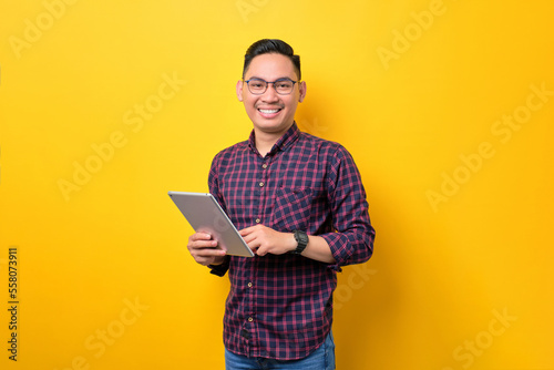 Smiling young Asian man in glasses holding digital tablet and looking at camera isolated over yellow background
