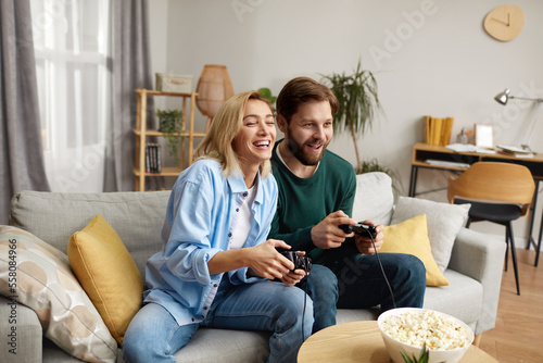 Happy Couple Playing Video Games. Boyfriend And Girlfriend Sitting On Couch In Living Room Enjoying Playing Video Games And Spending Time Together. Enjoying Moment On Weekend Concept