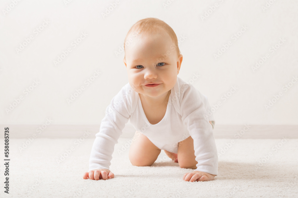 Happy smiling adorable baby boy in white bodysuit crawling on knee and arms on light beige home carpet. Posing and looking at camera. 5 to 6 months old infant development. Front view. Closeup. Happy smiling adorable baby boy in white bodysuit crawling on knee and arms on light beige home carpet. Posing and looking at camera. 5 to 6 months old infant development. Front view. Closeup.