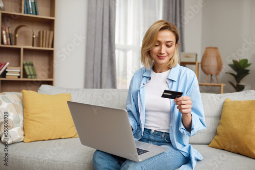 Positive Woman Holding Credit Card. Smiling Woman Paying Online, Using Laptop, Holding Plastic Credit Card, Sitting On Couch At Home. Young Female Shopping, Making Secure Internet Payment 
