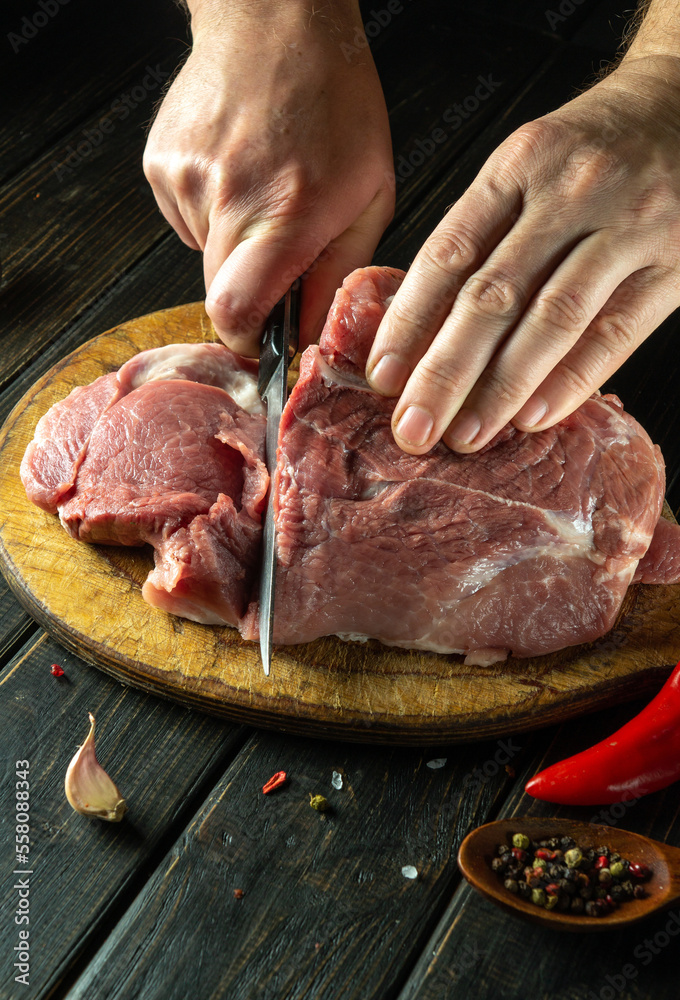 The cook cuts a raw beef steak with a knife on a wooden cutting board. The concept of the process of preparing a meat dish for lunch with aromatic spices.