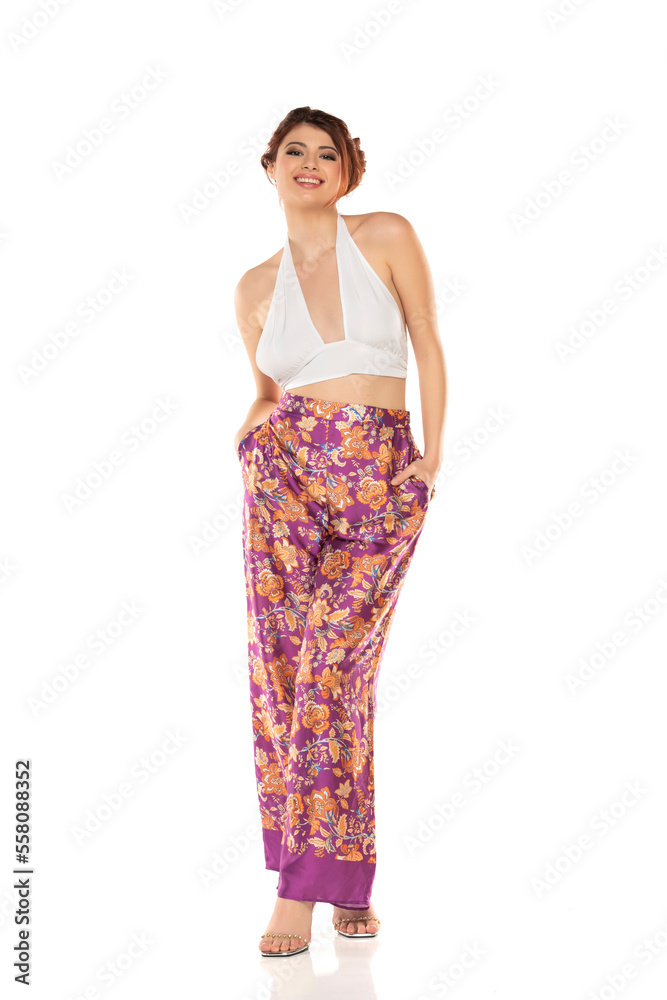 Fashion style studio portrait of beautiful young woman. Floral pattern wide leg pants and white sleevles shirt. Model standing and posing against white background