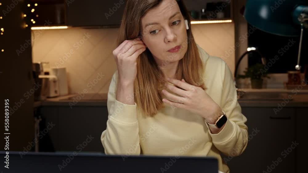 Woman wearing earphones sitting at table in dark room and using laptop. Working late from home office. Female freelancer works in home office at night, use laptop Woman wearing earphones sitting at table in dark room and using laptop. Working late from home office. Female freelancer works in home office at night, use laptop