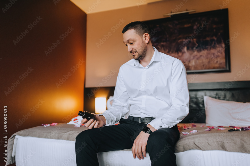 A stylish pensive man in a white shirt, a bearded groom sits on a bed in a room in the morning and looks at an expensive perfume. Close-up wedding photography, portrait.