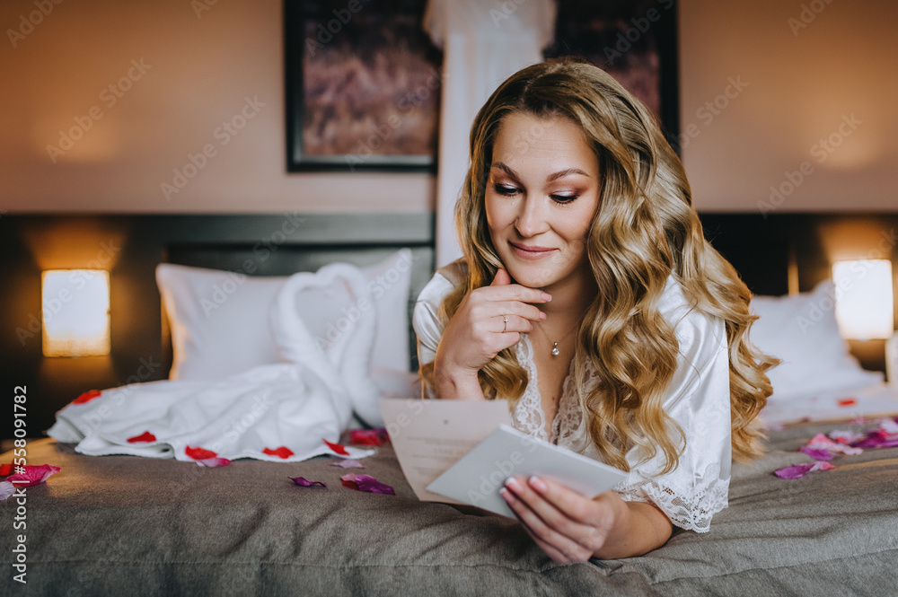 A beautiful smiling curly blonde bride in lingerie, a dressing gown lies in the morning in a room on a bed in rose petals, a bedroom, reading an oath from a paper envelope. wedding photography. A beautiful smiling curly blonde bride in lingerie, a dressing gown lies in the morning in a room on a bed in rose petals, a bedroom, reading an oath from a paper envelope. wedding photography.
