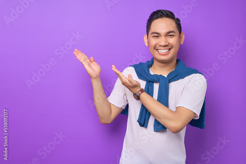 Smiling young Asian man 20s wearing white t-shirt points his hands arms aside indicate on workspace area isolated on purple background. People lifestyle concept