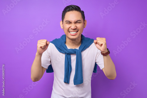 фотографія overjoyed and excited young Asian man 20s wearing white t-shirt doing a winner gesture celebrate clenching fists say yes isolated on purple background