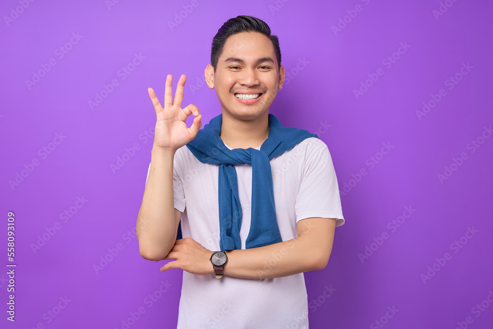 Smiling satisfied and cool young Asian man 20s wearing white t-shirt casual clothes showing okay ok gesture isolated on purple background. People lifestyle concept