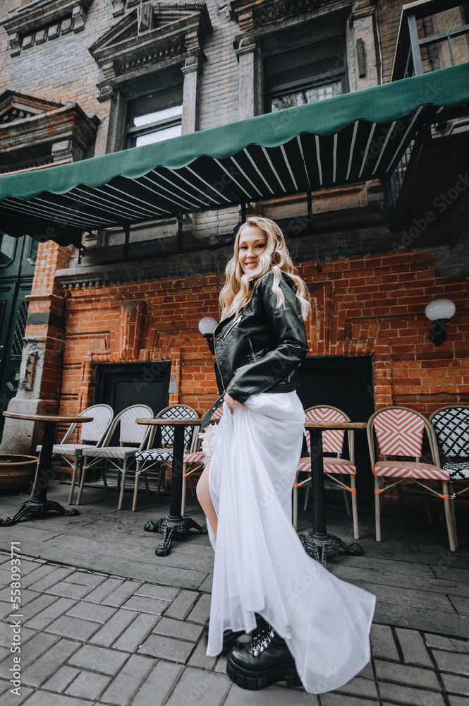 A beautiful curly bride, a biker, a rock lover in a black leather jacket, a white dress with a bouquet, walks in the city against the backdrop of a brick wall. Wedding photography, portrait.