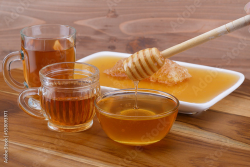 a bowl of honey with honey dipper, glass cup of tea, and honeycomb on wooden table