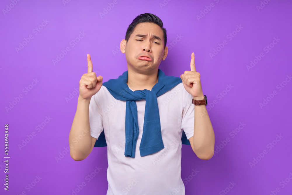 sad young Asian man wearing white t-shirt pointing fingers up at copy space isolated over purple background. people lifestyle concept sad young Asian man wearing white t-shirt pointing fingers up at copy space isolated over purple background. people lifestyle concept