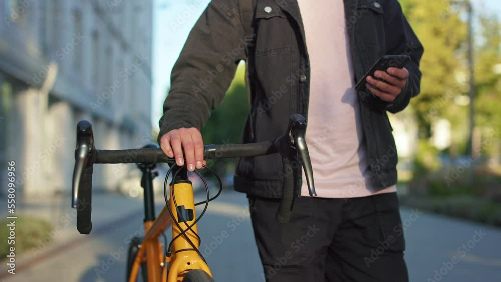 Close up of young male walking with bicycle, holding smartphone, using. Man wearing casual clothes, riding yellow bike outdoors in summer. Concept of modern urban lifestyle. Close up of young male walking with bicycle, holding smartphone, using. Man wearing casual clothes, riding yellow bike outdoors in summer. Concept of modern urban lifestyle.