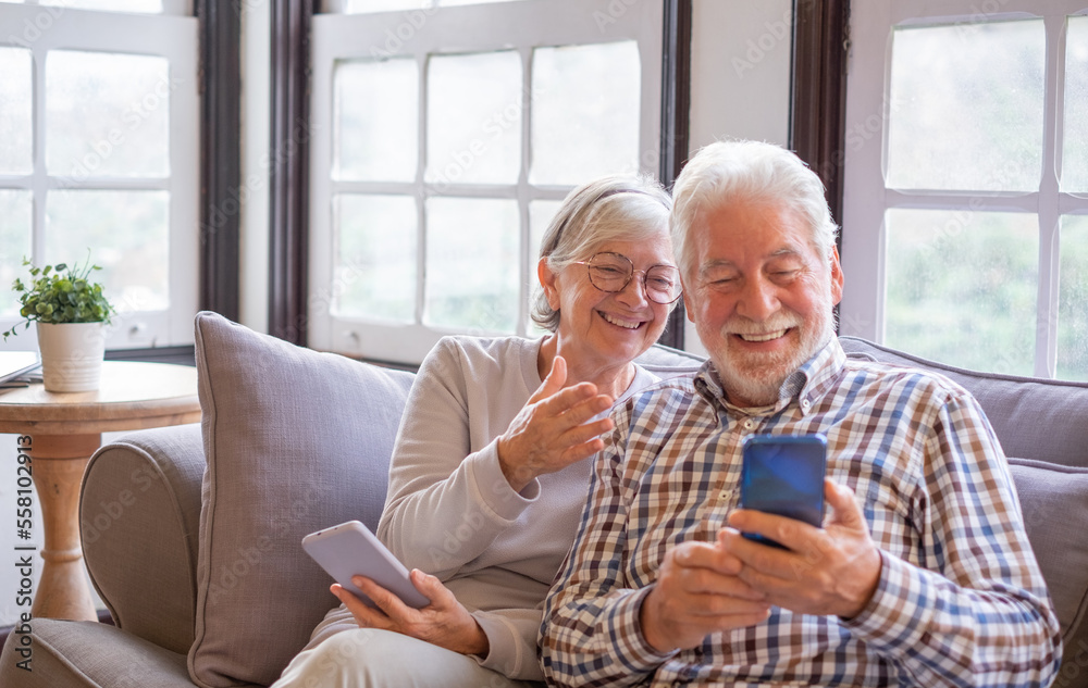 Portrait of cheerful senior couple while sitting together on sofa laughing. Elderly happy couple relaxing while using smartphones Portrait of cheerful senior couple while sitting together on sofa laughing. Elderly happy couple relaxing while using smartphones