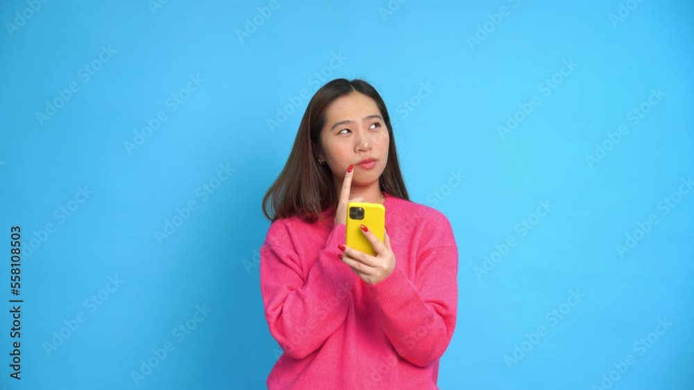 Thoughtful young Asian female in casual clothes looking at camera and touching lips while browsing mobile phone against blue background Thoughtful young Asian female in casual clothes looking at camera and touching lips while browsing mobile phone against blue background
