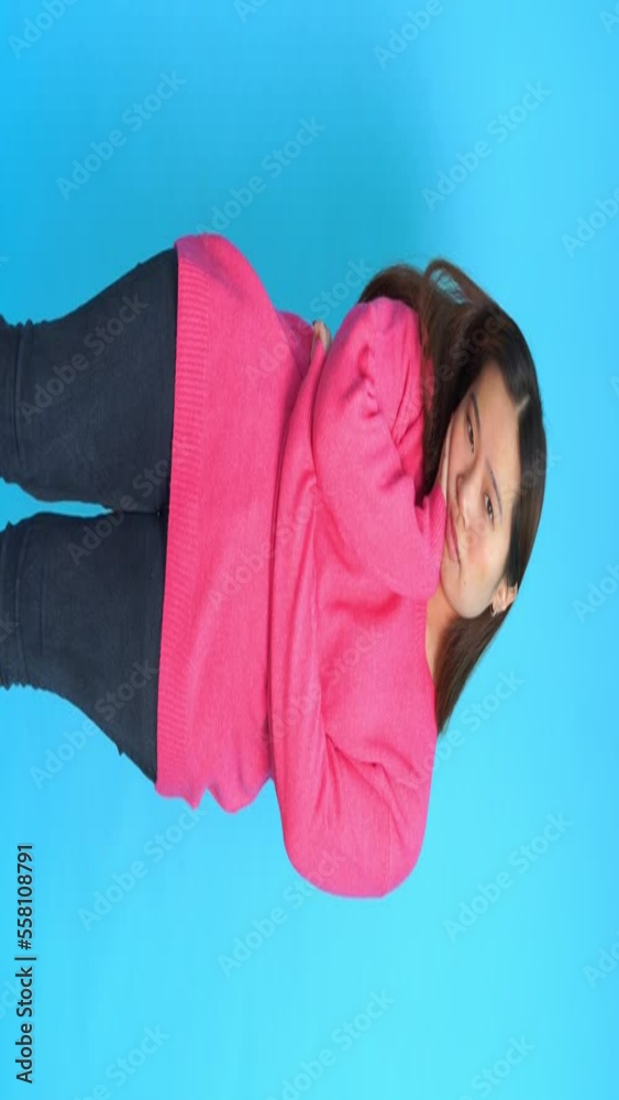 Positive young female in pink sweater looking at camera with astonished face expression while standing on blue background in studio Positive young female in pink sweater looking at camera with astonished face expression while standing on blue background in studio